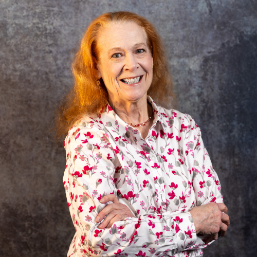 A woman with long strawberry-blonde hair smiles with her arms crossed. She is wearing a white button-down shirt featuring a pattern of pink and grey flowers. The background is a grey, textured studio wall.