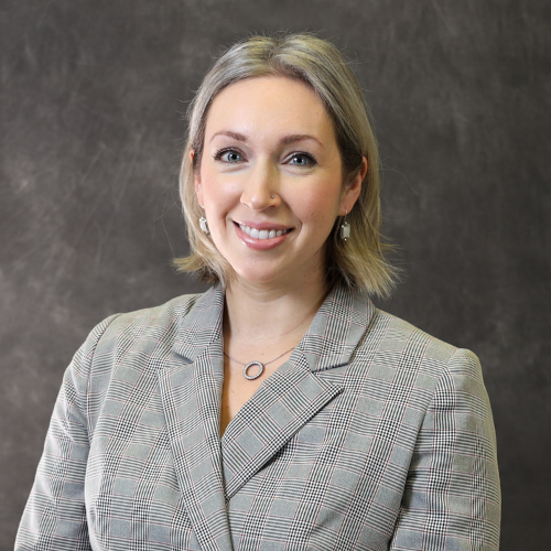 Nicole Watson, a woman with blonde hair wearing a grey plaid blazer and a silver circle necklace, smiling against a dark grey studio backdrop.