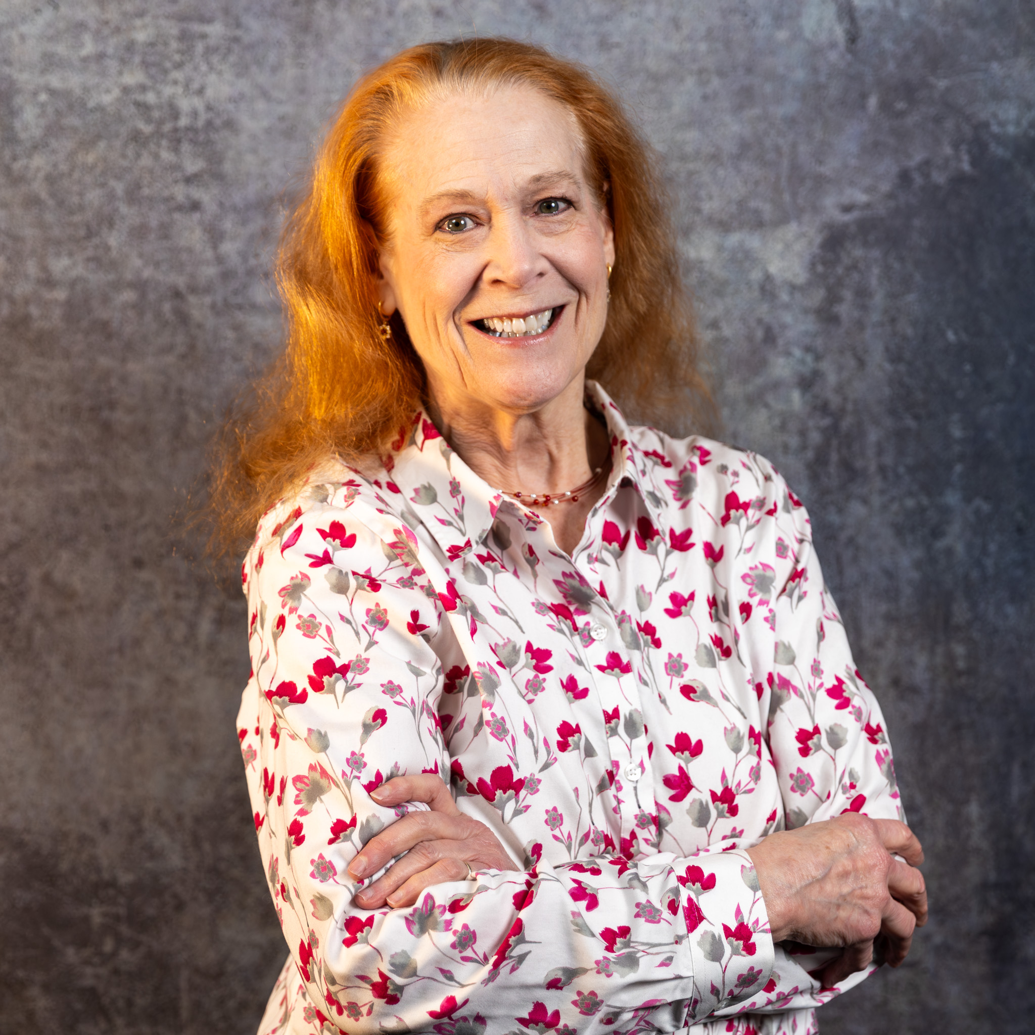 A woman with long strawberry-blonde hair smiles with her arms crossed. She is wearing a white button-down shirt featuring a pattern of pink and grey flowers. The background is a grey, textured studio wall.