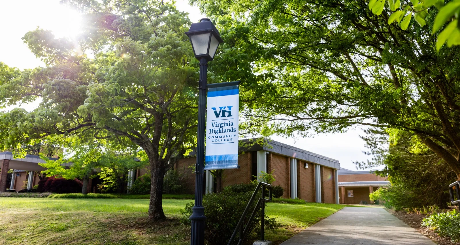 A sunlit pathway on the Virginia Highlands Community College campus, lined with lush green trees and brick buildings. In the foreground, a black lamppost features a blue and white VHCC banner.