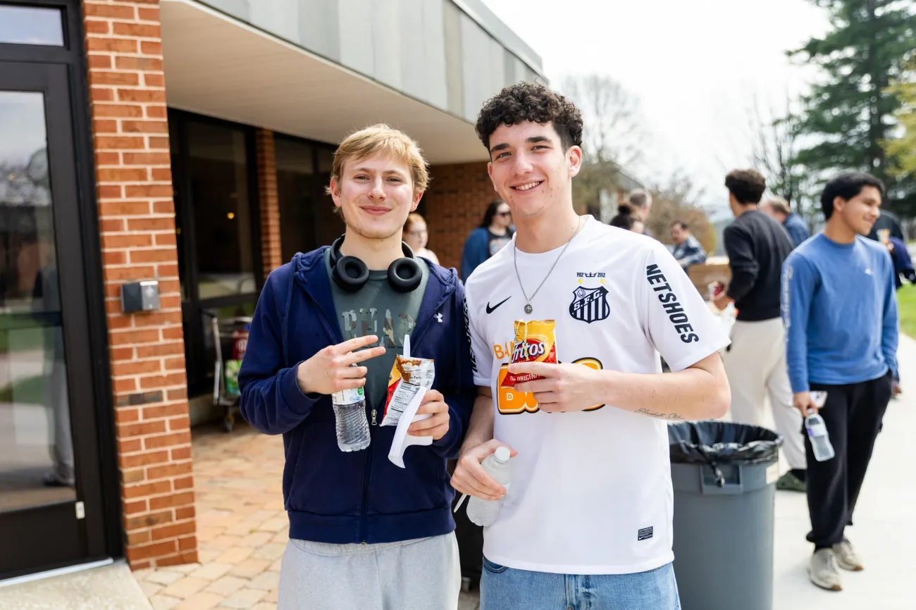 Two smiling male students stand together outdoors during a campus event, holding snacks and water bottles. The student on the left wears a blue zip-up hoodie and headphones around his neck, while the student on the right wears a white Santos FC soccer jersey. Other students and a brick building are visible in the background.