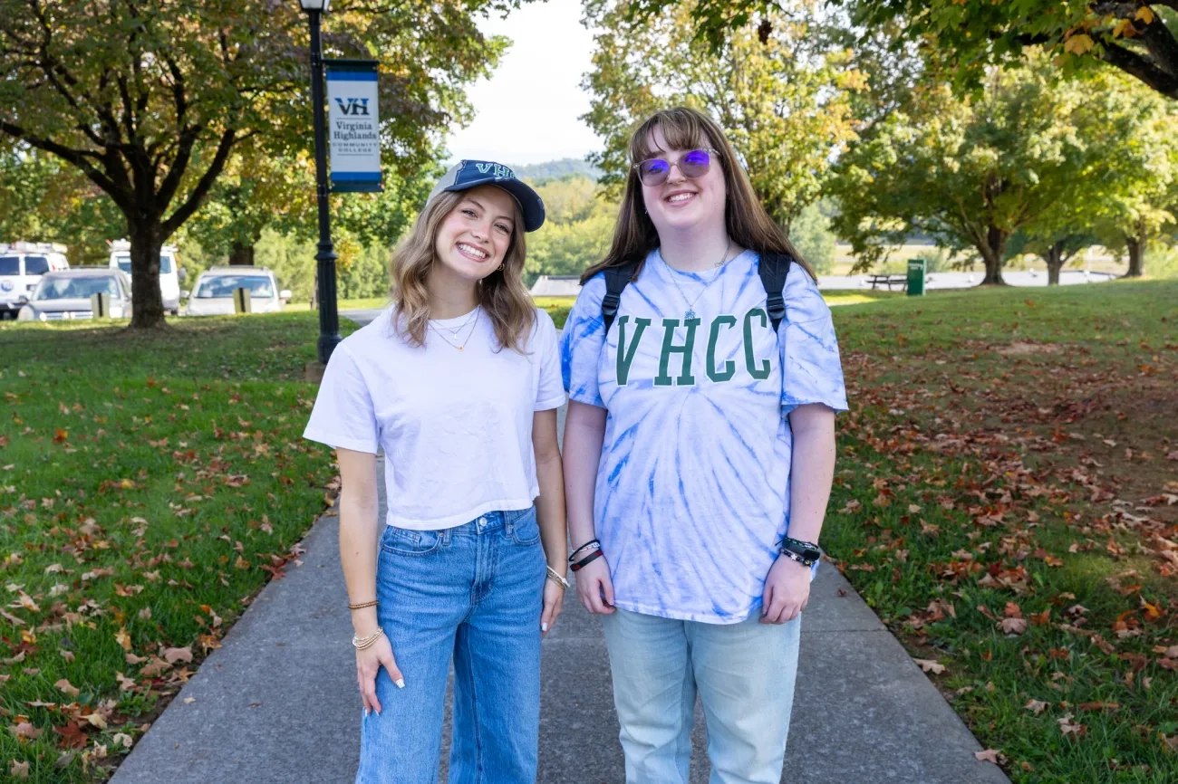 Two smiling female students stand side-by-side on a paved campus walkway at Virginia Highlands Community College. The student on the left wears a white t-shirt, jeans, and a VHCC cap, while the student on the right wears a blue tie-dye VHCC shirt and glasses, with green trees and a campus banner in the background.