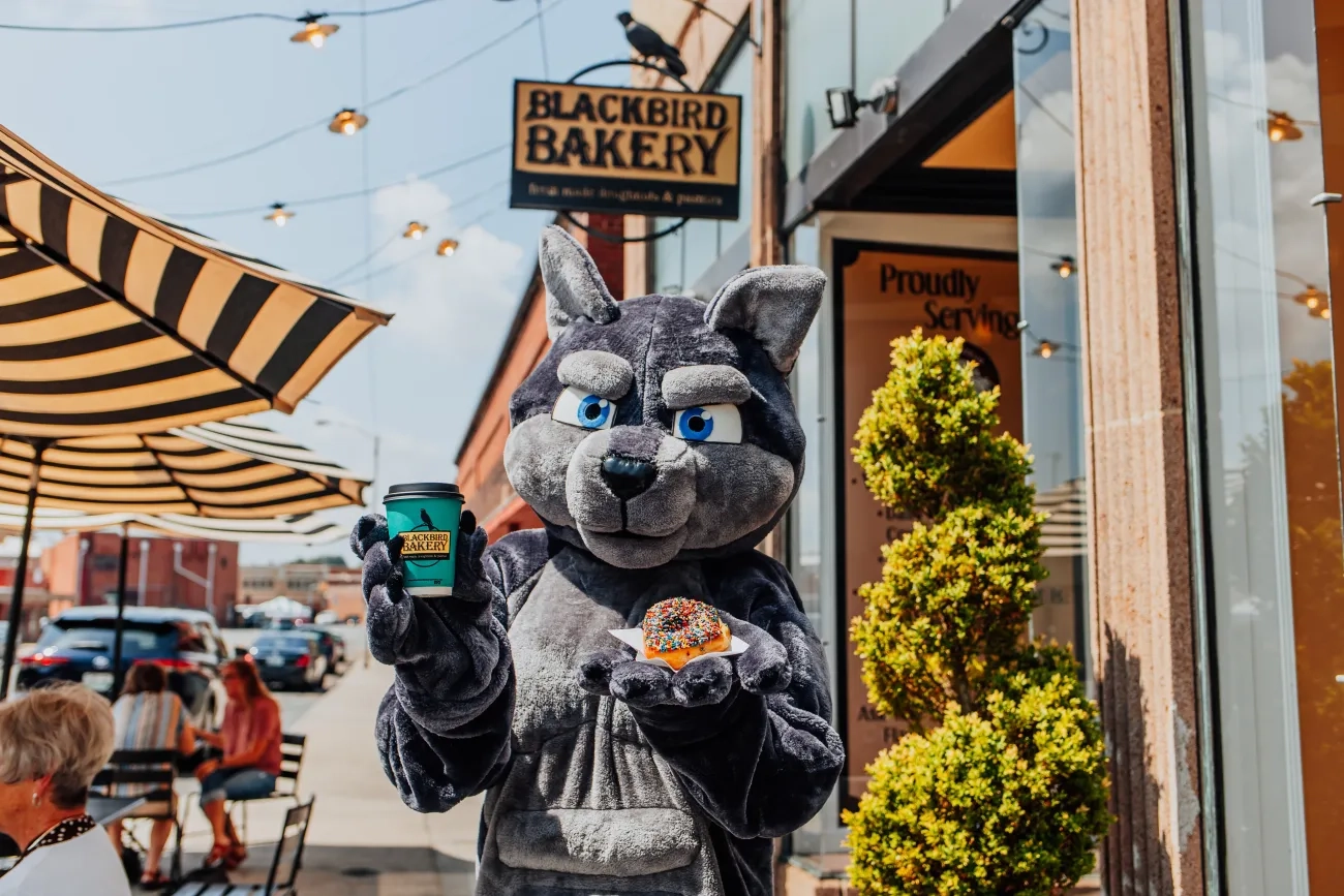 VHCC wolf mascot holding a donut and coffee in front of discount partner, Blackbird Bakery