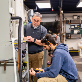 An HVAC instructor in a grey polo shirt and glasses stands over a student kneeling in an active workshop. The student is wearing a blue hoodie and safety glasses while using a digital multimeter to test wires inside an open heating and cooling unit.