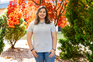 Outdoor portrait of VHCC alumna Jaquelin Cruz smiling warmly, wearing a grey and white striped t-shirt and blue jeans, standing in front of vibrant red and green autumn trees.