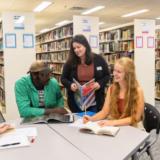 Four students studying together at a gray modular table inside a campus library surrounded by rows of bookshelves. Two students are seated reviewing open textbooks and notebooks, while a female staff member stands beside them holding a book featuring an American flag design.