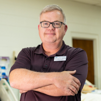 Professional headshot of Aaron Osborne, a man with glasses and short grey hair, smiling with arms crossed. He is wearing a maroon polo shirt and a Virginia Highlands name tag against a blurred classroom or medical training lab background.