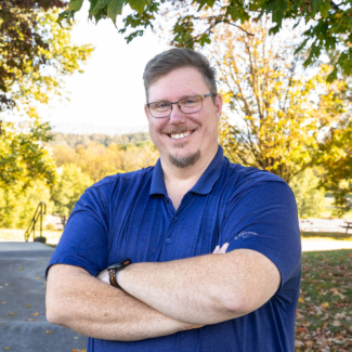 Professional headshot of Adam Brock, a smiling man with glasses and a goatee, wearing a navy blue polo shirt, standing outside under the shade of autumn trees.