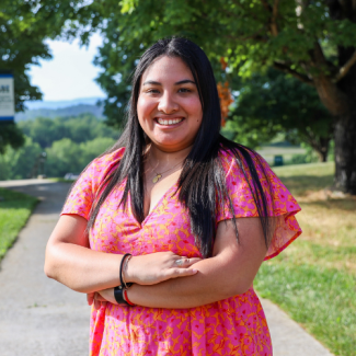 Headshot of Alexandra Centeno wearing a pink and orange floral dress, smiling with arms crossed in an outdoor park setting.