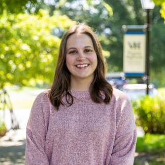 Amanda Fleenor smiling outdoors, wearing a pink heathered sweater and a thin gold necklace, with green trees and a blurred campus sign in the background.