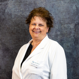 A woman with short, dark, styled hair smiles warmly against a grey textured background. She is wearing a professional white lab coat over a black shirt, with a nametag identifying her as Amy Hampton, Associate Professor of Nursing.