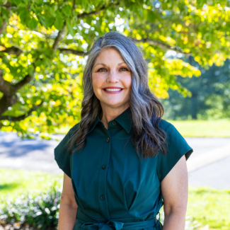 Headshot of Andi Fultz, a smiling woman with wavy grey-and-dark hair wearing a dark green short-sleeved shirt outdoors.