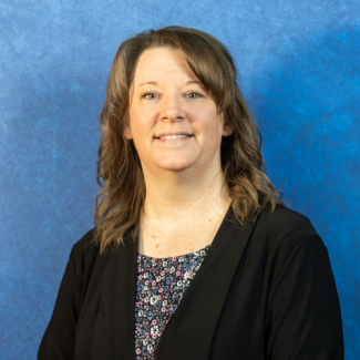 A professional headshot of Andrea Repass, a woman with shoulder-length wavy brown hair, smiling and wearing a black cardigan over a floral print blouse against a textured blue background.