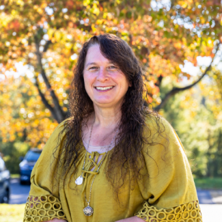 Angela Melvin smiling, wearing a mustard-yellow top and necklaces outdoors in front of autumn trees.