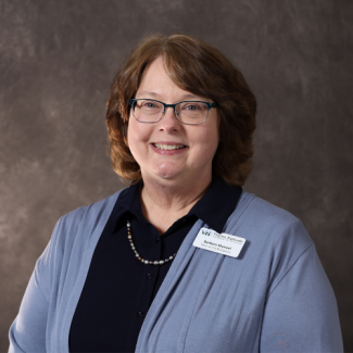 Barbara Manuel wearing glasses, a navy shirt, and a light blue cardigan. She has a name tag that identifies her as Dean of Arts & Sciences.