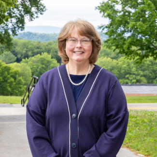 A smiling woman with shoulder-length light brown hair and glasses, wearing a dark blue top, a purple cardigan with white trim, and a pearl necklace. She is standing outdoors with a background of green hills and trees.