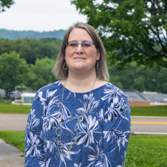 Headshot of Rebecca Kell smiling outdoors, wearing glasses, a blue and white floral blouse, and a silver necklace.