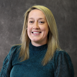Portrait of Bethaney Hilt, a woman with long blonde hair, wearing a dark teal textured blouse against a gray studio background.