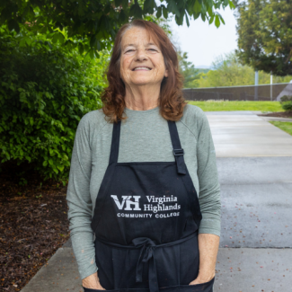 Headshot of Betty Duty wearing a gray long-sleeved shirt and a black Virginia Highlands Community College apron, smiling outdoors.