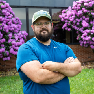 Brad Pennington, a man with a beard and glasses wearing a green VHCC baseball cap and a blue athletic shirt, standing with arms crossed.