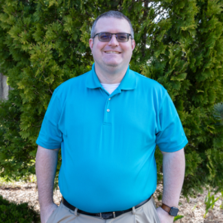 Brandon Hensley smiling outdoors in a bright blue polo shirt and glasses, standing in front of green shrubbery.