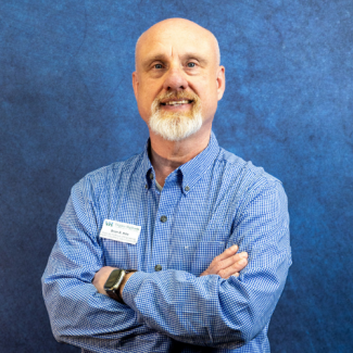 A professional headshot of Brian D. Hale, a man with a shaved head and a graying goatee, smiling with arms crossed. He wears a blue gingham shirt and a Virginia Highlands Community College name tag against a textured blue background.