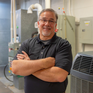 Bruce Olivo with a goatee and glasses, standing with arms crossed in a technical or mechanical room with HVAC equipment.