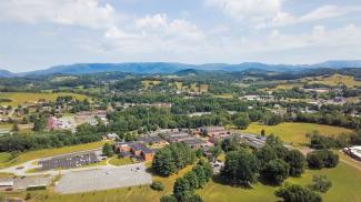 An aerial, high-angle drone photograph showing the Virginia Highlands Community College campus buildings surrounded by green trees, open fields, and parking lots, with rolling blue mountains visible under a partly cloudy sky in the distance.