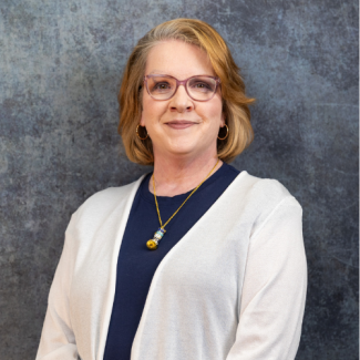 A woman with blonde hair and glasses smiles warmly for a portrait. She is wearing a teal blue blouse and a silver necklace with a circular pendant. The background is a grey, textured studio wall.