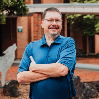 Headshot of Chris Church wearing glasses and a blue polo shirt, smiling with arms crossed in front of a brick building.