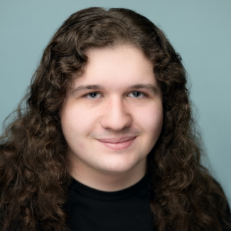 A close-up portrait of a young man with long, curly brown hair and a slight smile. He is wearing a black t-shirt against a solid, light blue-grey background.