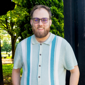 Colton Williams, a man with a beard and glasses, wearing a light blue and white vertical-striped polo shirt.