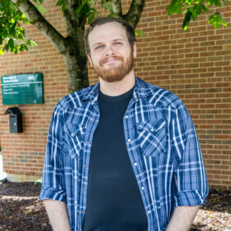 Headshot of Dallas Nelson, a man with a beard and short brown hair wearing a blue and white plaid shirt over a black t-shirt, standing in front of a brick building.