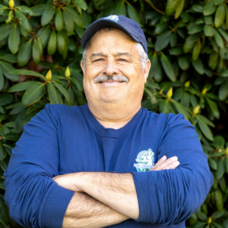 A man with a grey mustache and a blue baseball cap smiles with his arms crossed. He is wearing a long-sleeved navy blue shirt with a small mascot logo on the chest. The background consists of green leafy rhododendron bushes.