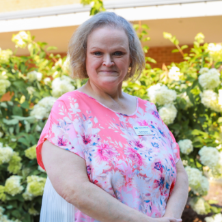 Headshot of Debbie Nelson, a woman with short, wavy blonde hair wearing a pink and white floral patterned blouse, standing in front of white flowering bushes.