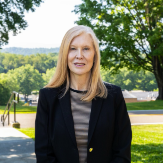 Deborah McCroskey smiling outdoors, wearing a black blazer over a black-and-beige horizontally striped top, with green trees and a hillside in the background.