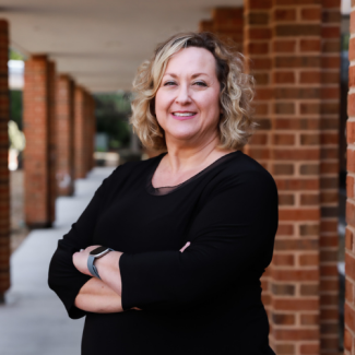 Headshot of Donna Corns wearing a black top, smiling with arms crossed in a brick-pillared outdoor walkway.