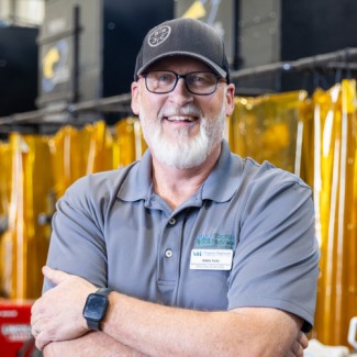 A smiling man with a white beard, glasses, and a black baseball cap poses with his arms crossed in an industrial setting. He wears a grey polo shirt with a "Virginia Highlands Community College" logo and a nametag identifying him as Eddie Fultz. Yellow welding curtains and industrial equipment are visible in the background.