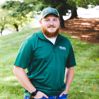 Headshot of Ein Denti wearing a green Virginia Highlands Community College polo and a VHCC baseball cap, smiling outdoors.