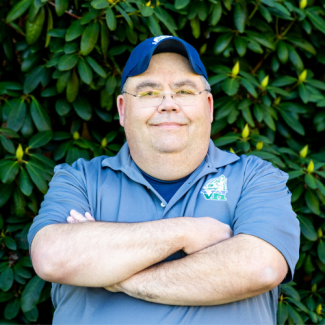 A man with glasses and a blue baseball cap smiles with his arms crossed in front of a green leafy bush. He is wearing a grey polo shirt with a small green and white mascot logo on the chest.