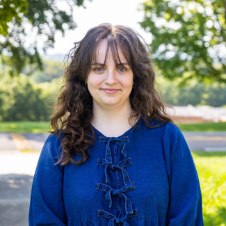 A outdoor portrait of Caitlin Manuel smiling against a soft-focused green background. She has shoulder-length, wavy brown hair with curtain bangs and wears a blue denim top with front bow details.