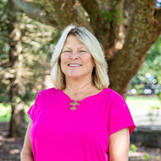 An outdoor headshot of a smiling woman with shoulder-length blonde hair, wearing a bright pink short-sleeve top with a decorative metal ring neckline, framed by trees.