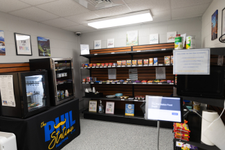 Wide angle view of a food pantry room featuring stocked food shelves, mini-refrigerators, a microwave, motivational signs, and a tablet check-in station in the foreground.