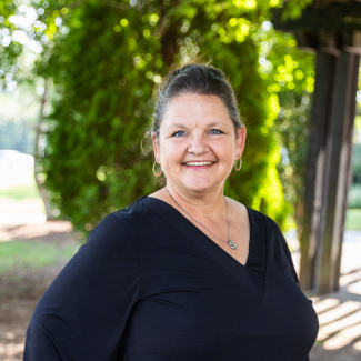 Close-up headshot of Sharon Rowe smiling outdoors, wearing hoop earrings and a black v-neck shirt.