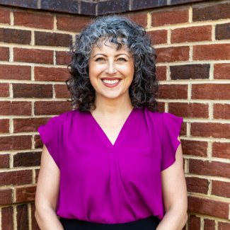Professional headshot of Hannah Ingram, a woman with curly grey and dark hair, smiling and wearing a vibrant purple blouse against a red brick wall background.
