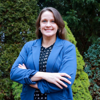 Hannah Mullins, smiling with arms crossed, wearing a blue blazer over a black and white patterned blouse.