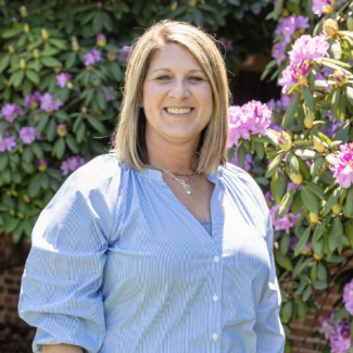 Headshot of Heather Perry, a woman with shoulder-length blonde hair, smiling in a light blue button-down shirt. She is standing outdoors in front of purple rhododendron flowers.