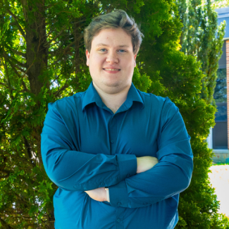 Headshot of Jared Adams wearing a teal button-down shirt, smiling with arms crossed in front of green evergreen trees.