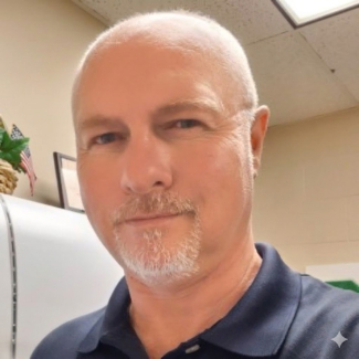 Headshot of Jeff Russell, a man with a shaved head and a graying goatee, looking directly at the camera with a neutral expression. He is wearing a navy blue polo shirt and is pictured in an office setting.
