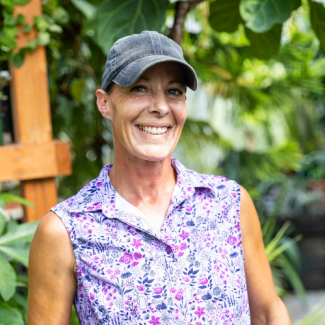 Headshot of Jennifer Dufresne, a smiling woman wearing a grey baseball cap and a purple floral sleeveless shirt outdoors.